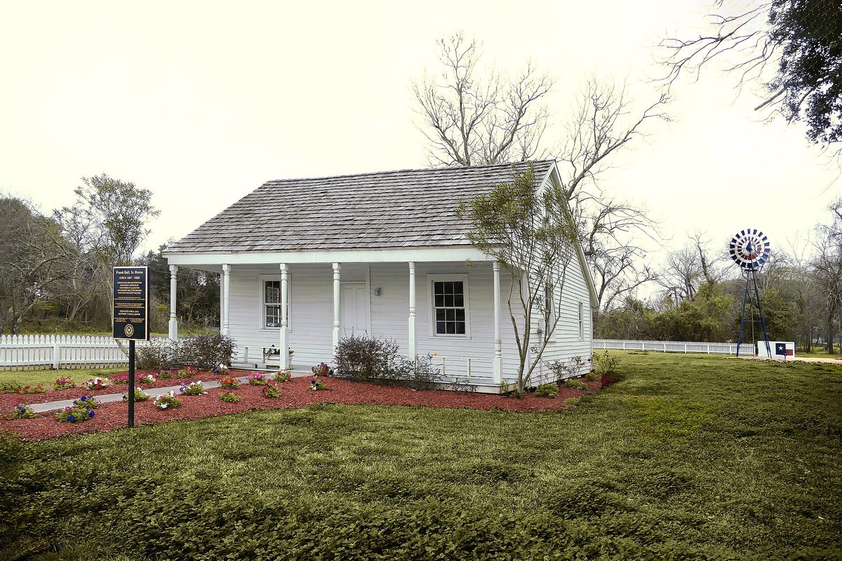 A small white cottage with a shingled roof and front porch in Texas City's Settlement Historic District, with a historical marker, flower beds and a windmill on the lawn