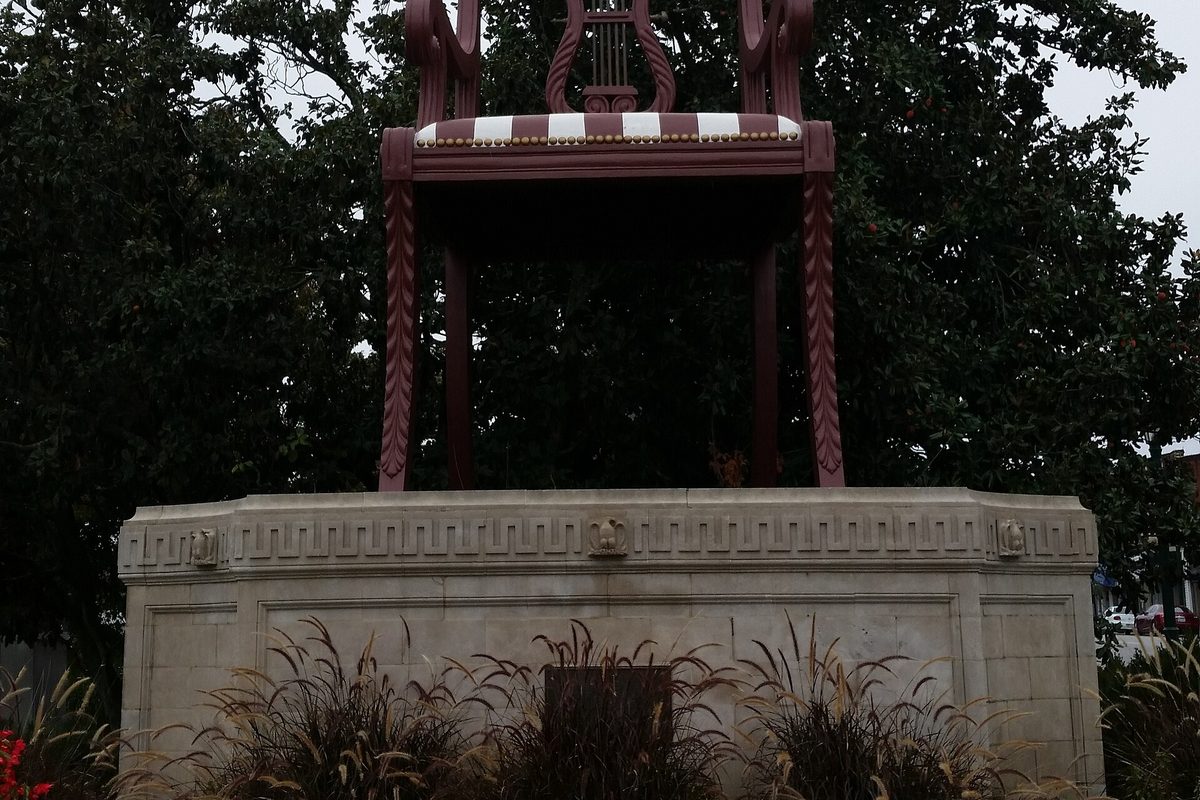 The Big Chair, a giant dark-red wooden chair with a striped seat, standing on a carved stone pedestal behind ornamental grasses, with trees in the background