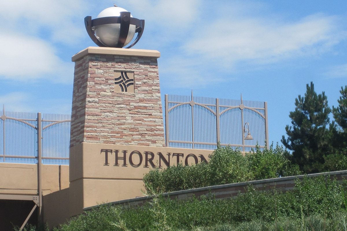 A stone pillar topped by a metal globe beside a tan wall with THORNTON in large letters, with a metal fence, pine trees and shrubs under a blue sky