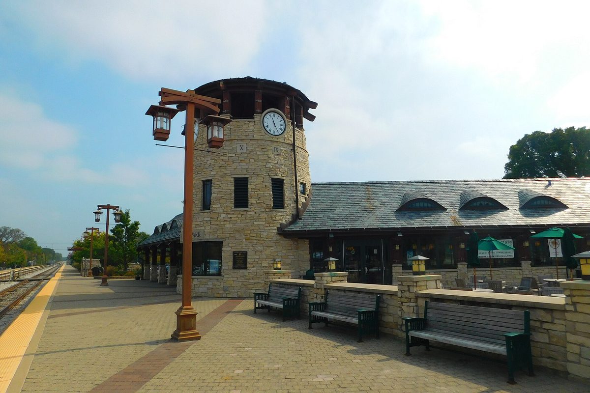 A Tinley Park Metra station with a round stone clock tower, a slate roof, lantern posts and green benches along a brick platform