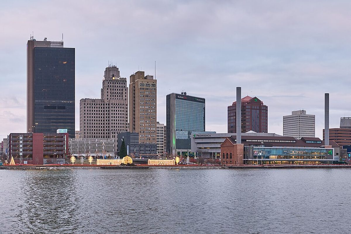 Downtown Toledo's skyline across the Maumee River on a gray evening, with holiday lights along the far bank and a brick smokestack among the office towers