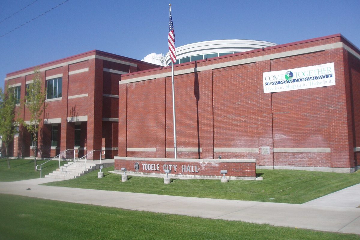 Tooele City Hall, a two-story red-brick building with a flagpole and a low brick sign reading Tooele City Hall, on a sunny day