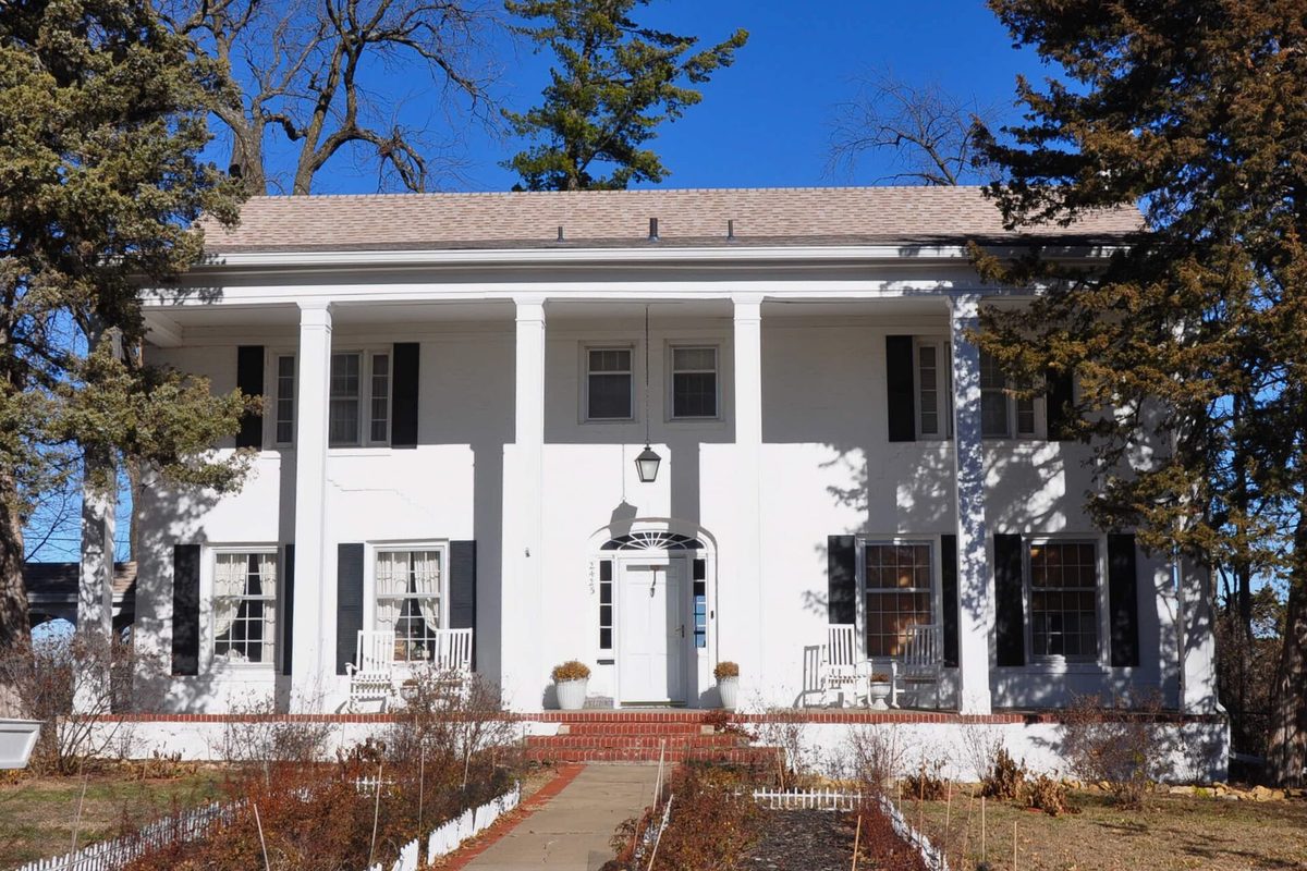 A white two-story house with square columns, black shutters and a brick front walk in Topeka's Country Club Residential Historic District, framed by evergreens