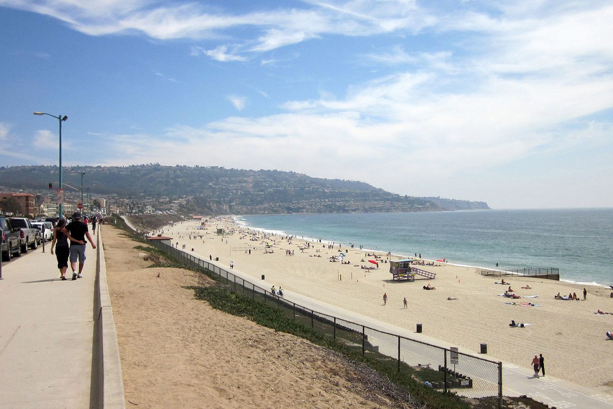 People walking along a bluff-top path above Torrance Beach, with sunbathers on the sand, a lifeguard tower and the Palos Verdes hills across the water