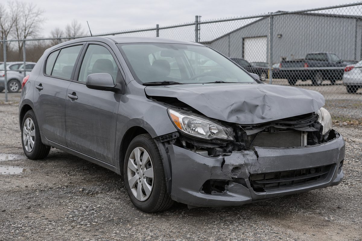 A grey hatchback with a crushed hood, broken headlight and torn front bumper parked on gravel by a chain-link fence
