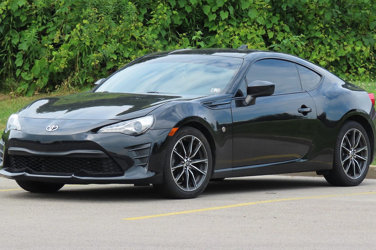 A black 2017 Toyota 86 coupe with dark multi-spoke alloy wheels, parked in a lot in front of thick green bushes