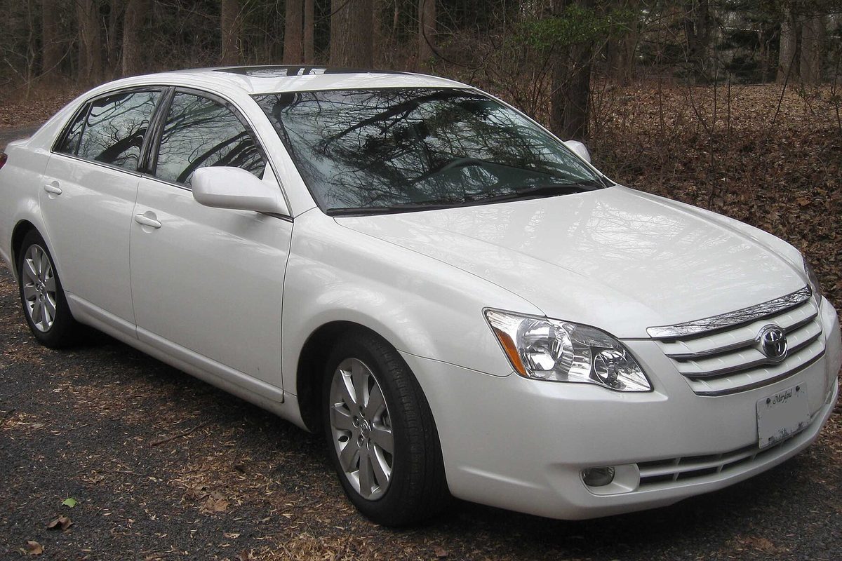 A white 2005-2007 Toyota Avalon sedan parked on a gravel drive among bare winter trees and fallen leaves