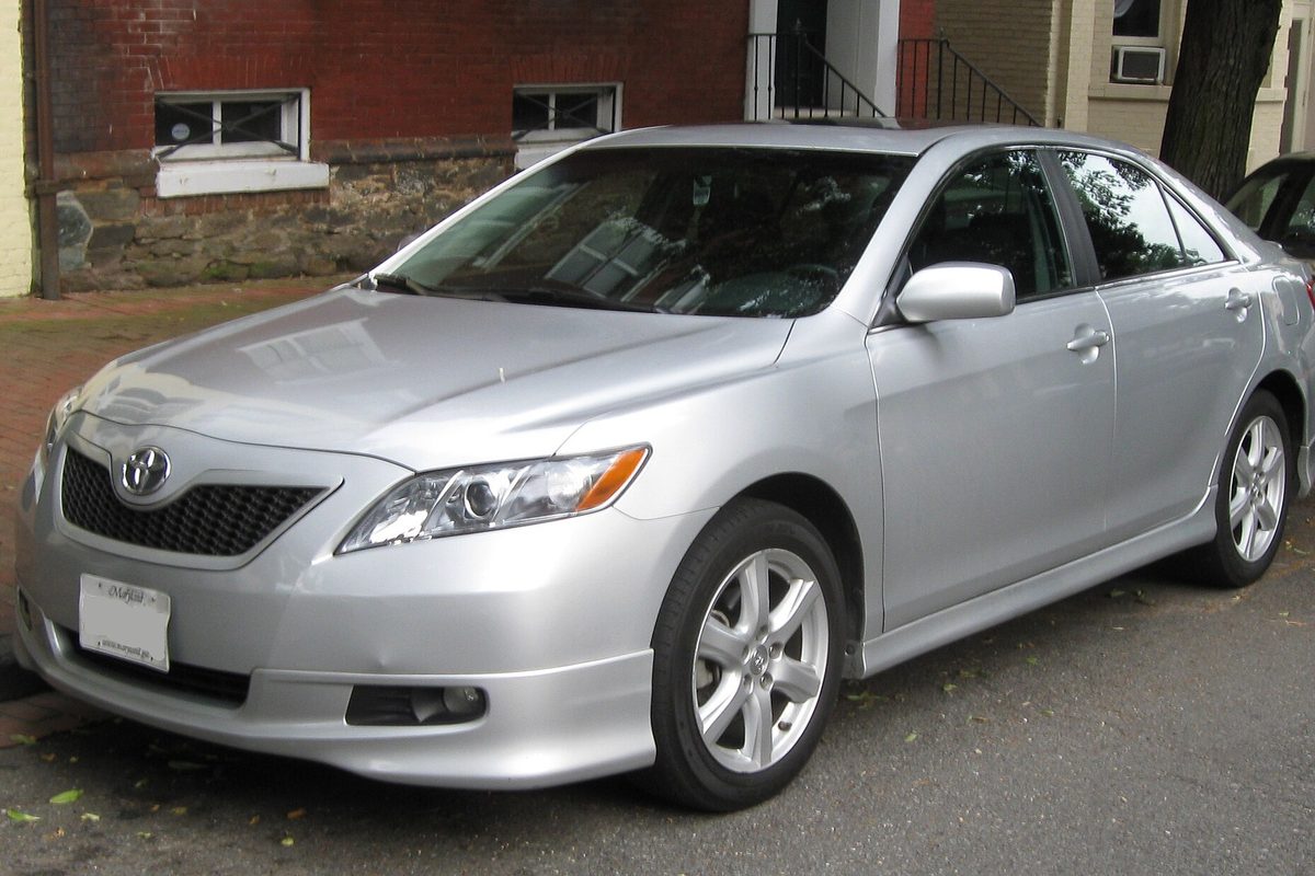 A silver 2007-2009 Toyota Camry SE parked on an asphalt street beside a brick sidewalk, in front of a red brick row house