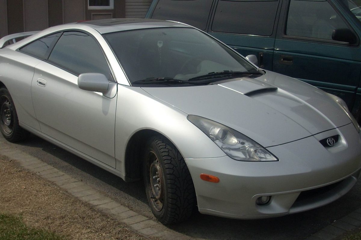 A silver 2000-2002 Toyota Celica GT-S liftback on black steel wheels and winter tires, parked in a driveway beside a green van