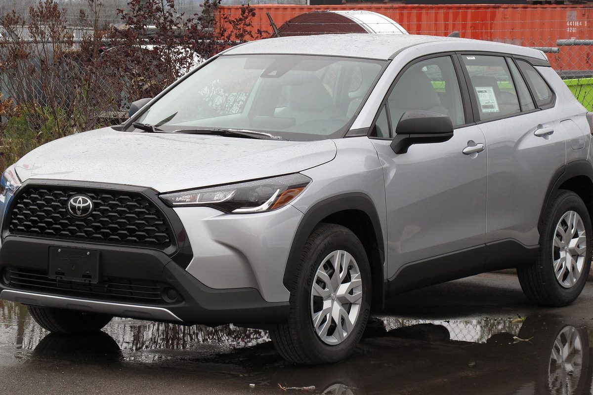 A silver 2022 Toyota Corolla Cross L on wet pavement, with red shipping containers and a leafless shrub behind it