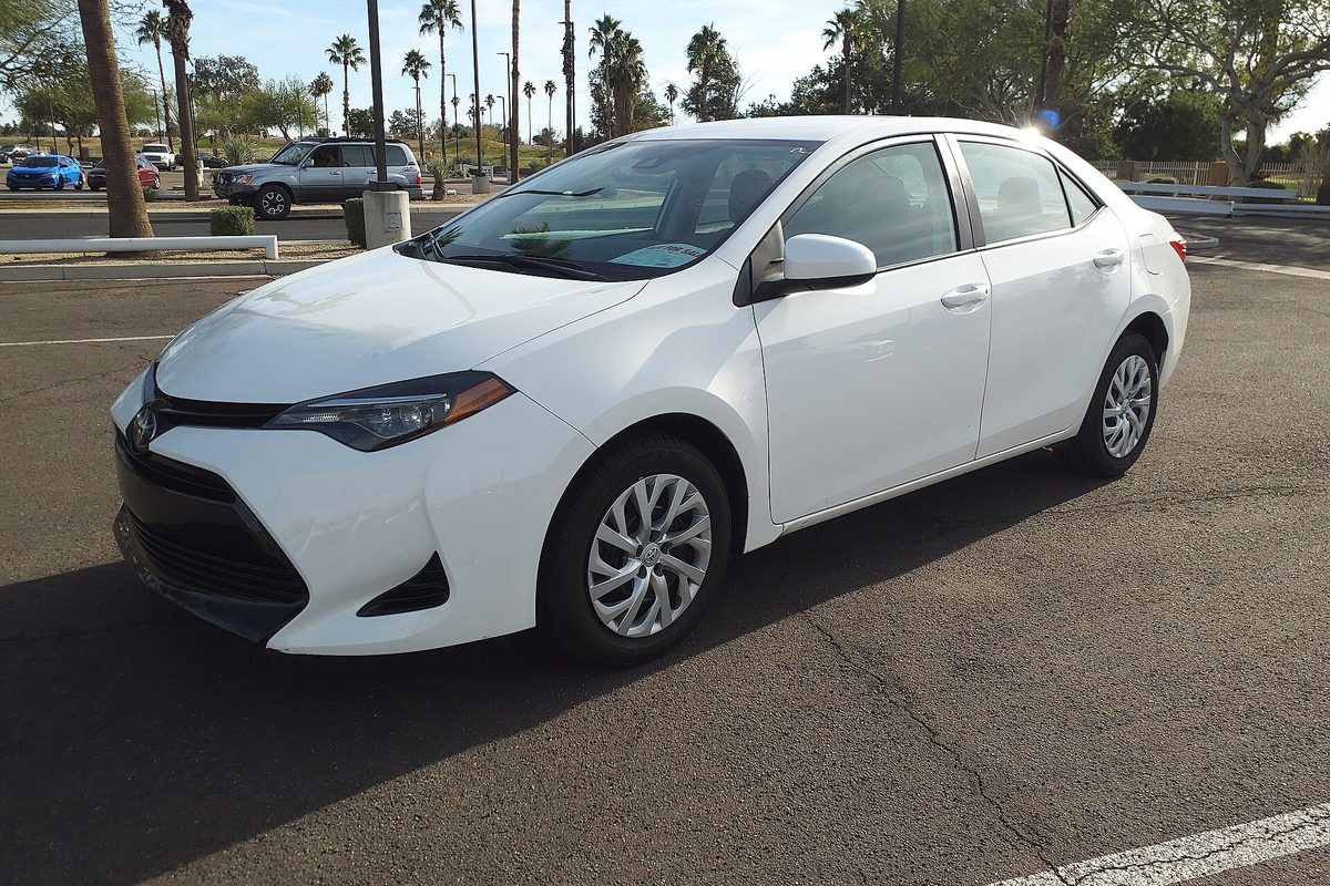 A white 2018 Toyota Corolla LE sedan in a sunny parking lot, with palm trees and parked cars behind it