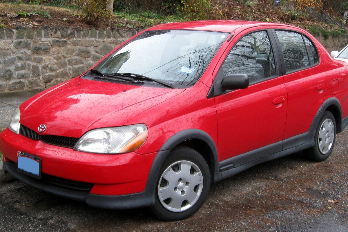 A red 2000-2002 Toyota Echo sedan with plastic wheel covers, parked on a wet street beside a stone wall