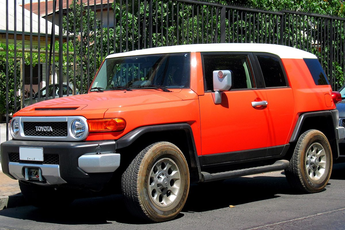 An orange Toyota FJ Cruiser with a white roof and mud on its tires, parked on a street beside a black iron fence