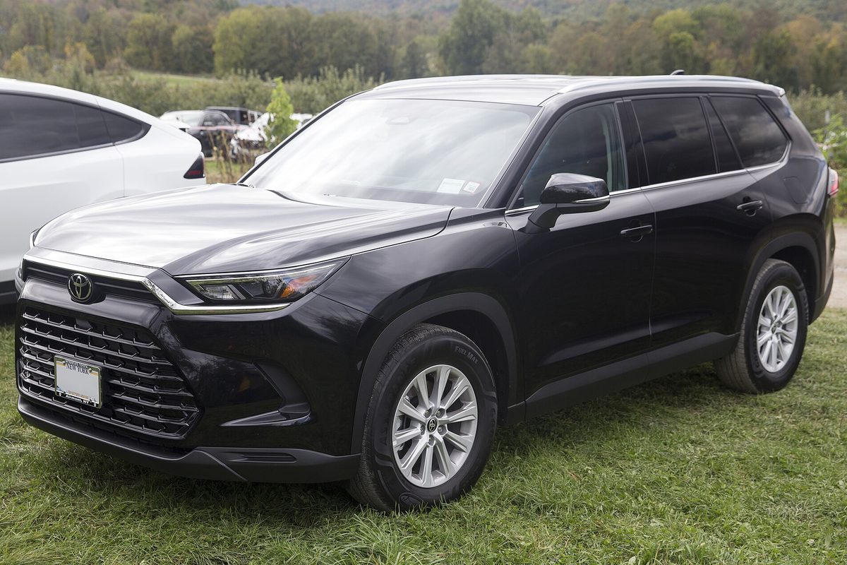 A black 2024 Toyota Grand Highlander XLE parked on grass beside other cars, with wooded hills behind