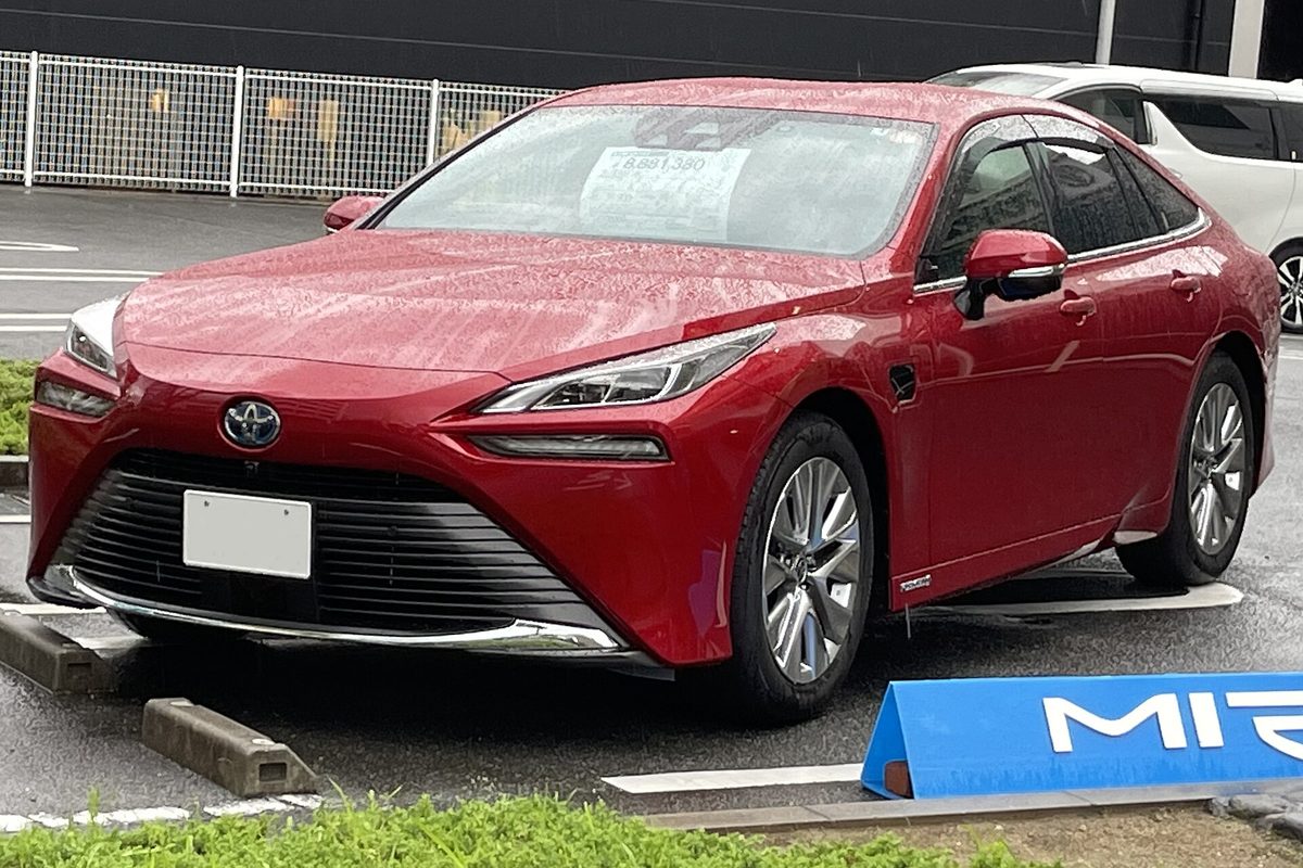 A red second-generation Toyota Mirai sedan on a rain-wet lot, with a blue Mirai sign in front of it