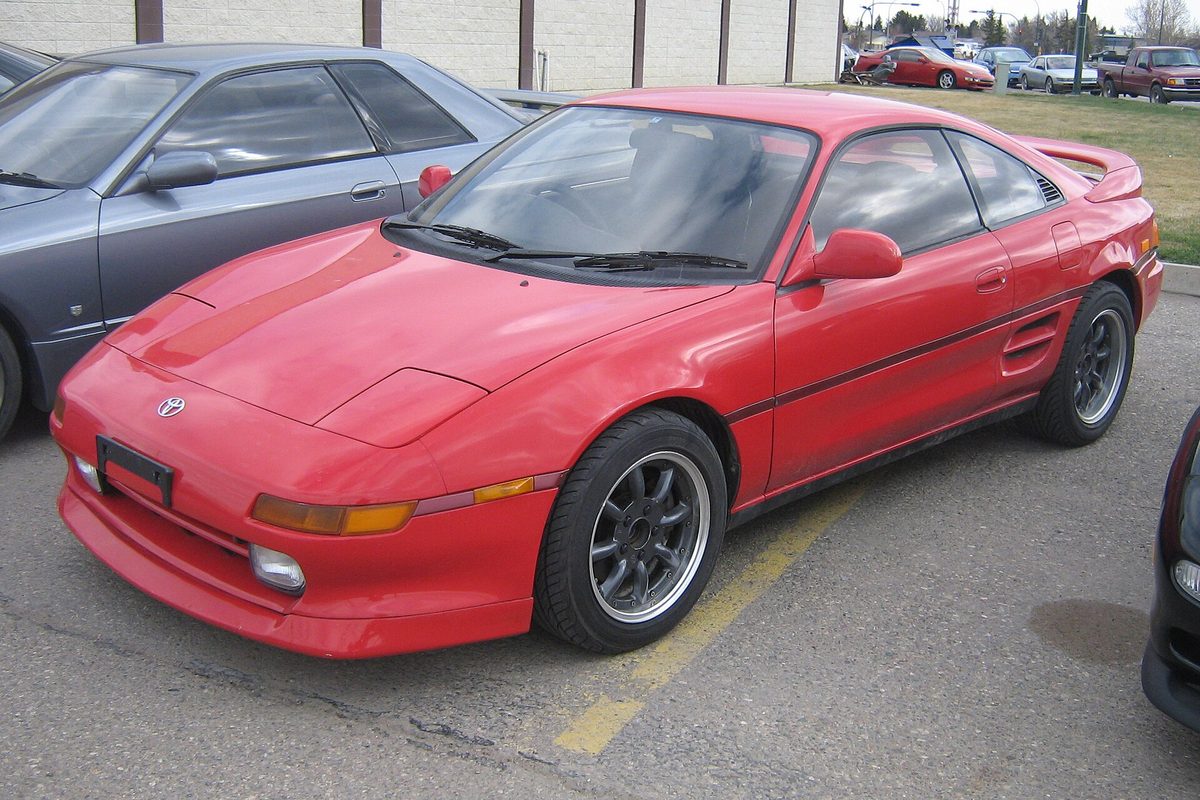 A red second-generation Toyota MR2 Turbo with dark Minilite-style wheels, parked in a lot beside a gray sedan