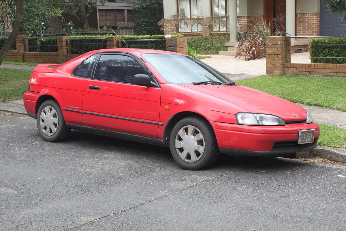 A red first-generation Toyota Paseo coupe parked at the curb of a quiet residential street, a brick garden wall behind it