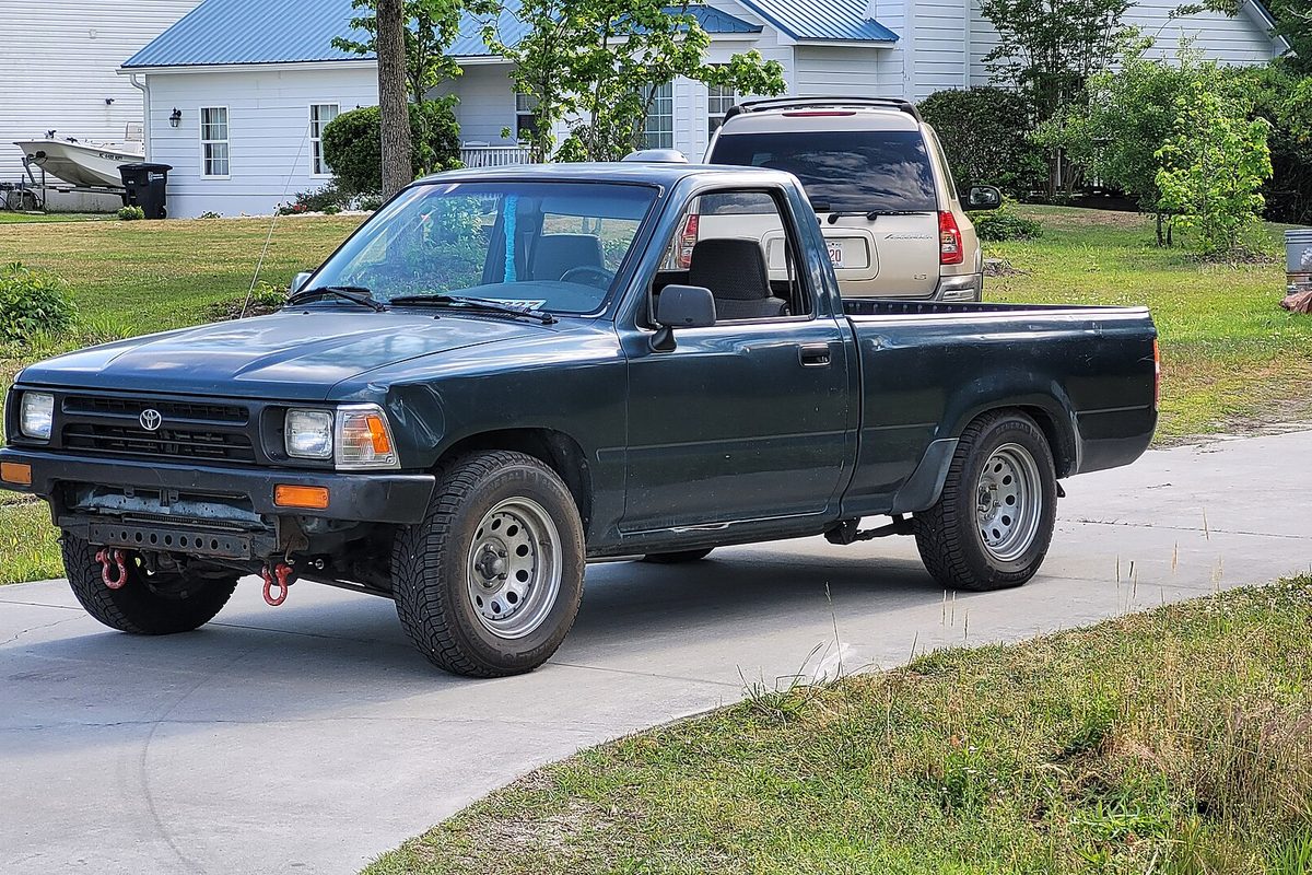A dark green 1994 Toyota Pickup regular cab on a concrete driveway, front bumper removed to show red tow hooks on the bare frame