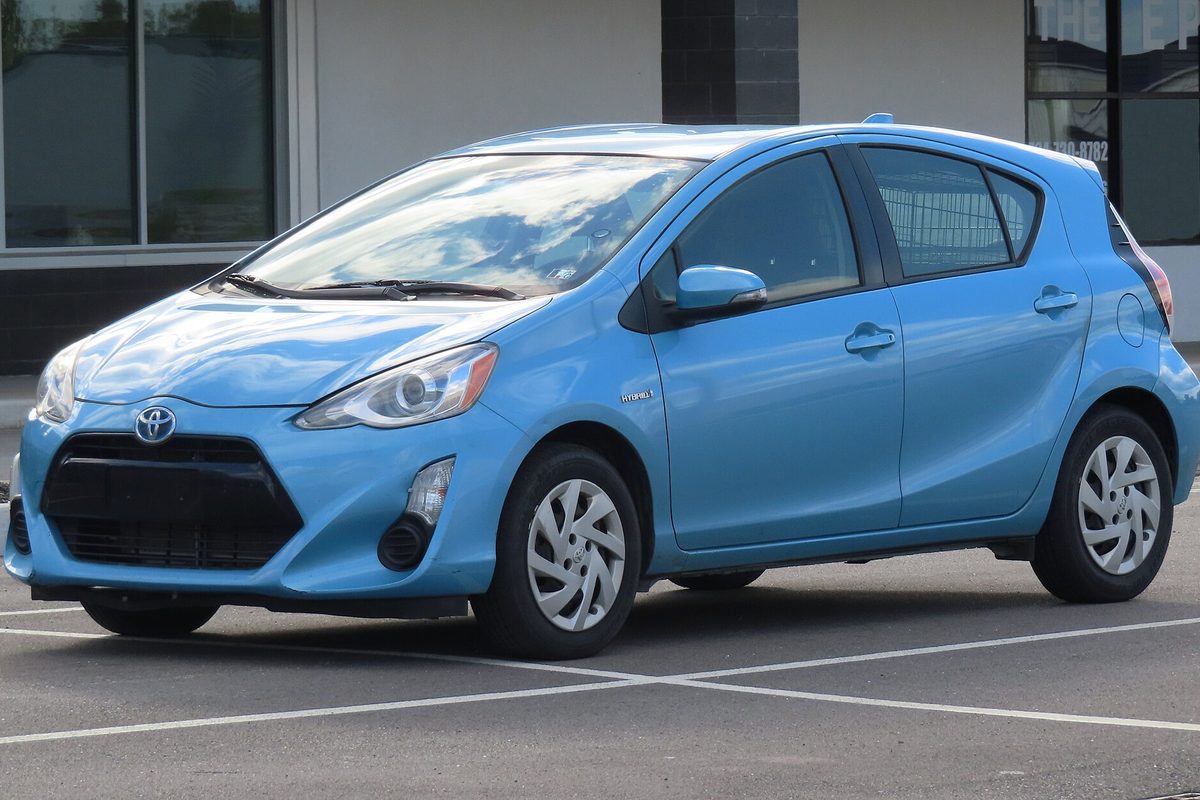 A light blue 2015 Toyota Prius c hatchback parked in an empty lot in front of a storefront