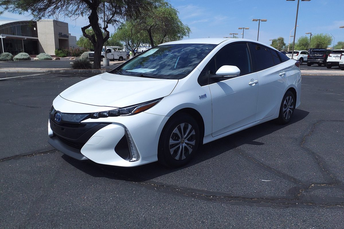 A white 2021 Toyota Prius Prime on a sunny parking lot, with desert trees, a work truck and parked SUVs behind it