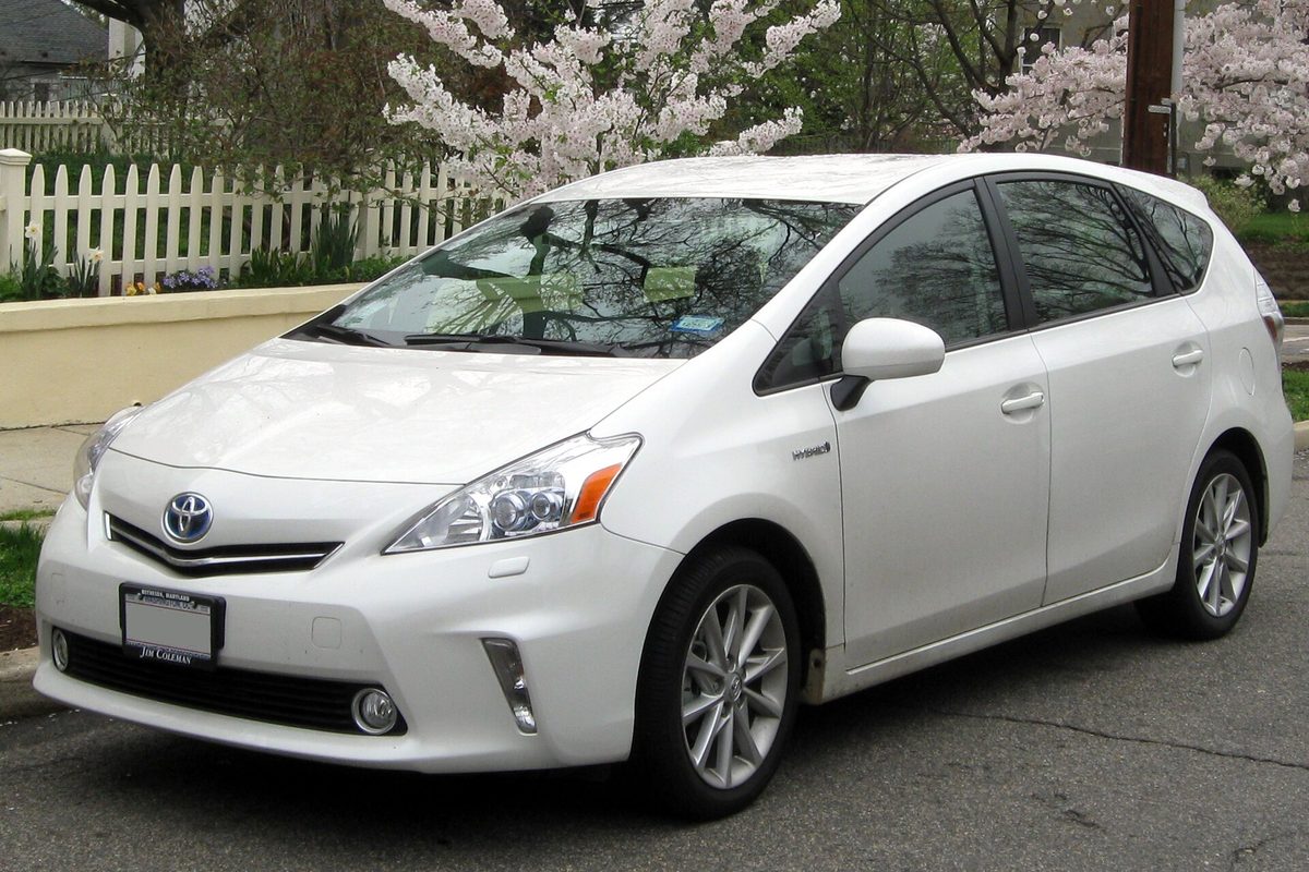 A white 2012 Toyota Prius v wagon parked on a street in front of a white picket fence and trees in blossom