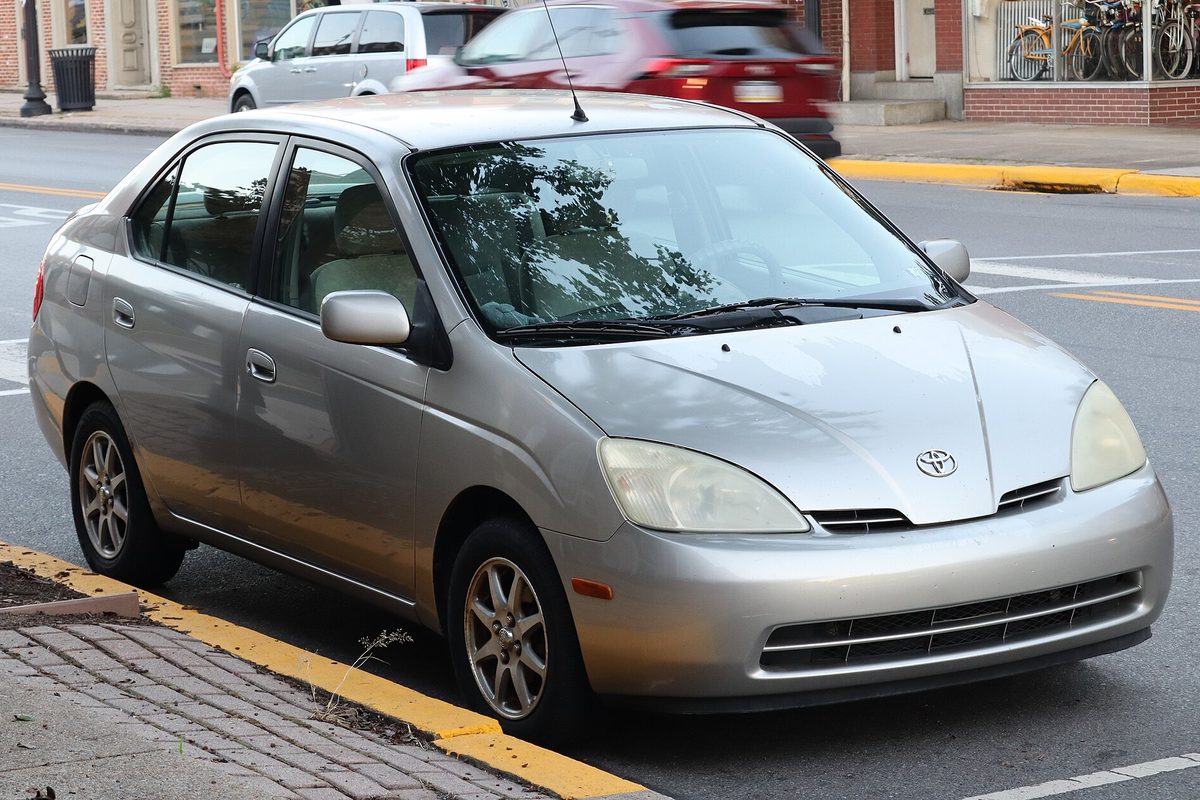 A silver first-generation 2003 Toyota Prius sedan parked at a yellow-painted curb beside a brick sidewalk