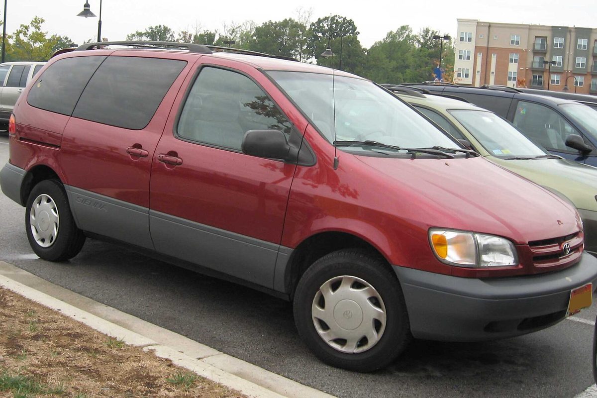 A dark red first-generation Toyota Sienna minivan with gray lower trim and plastic wheel covers, parked at a curb in a parking lot