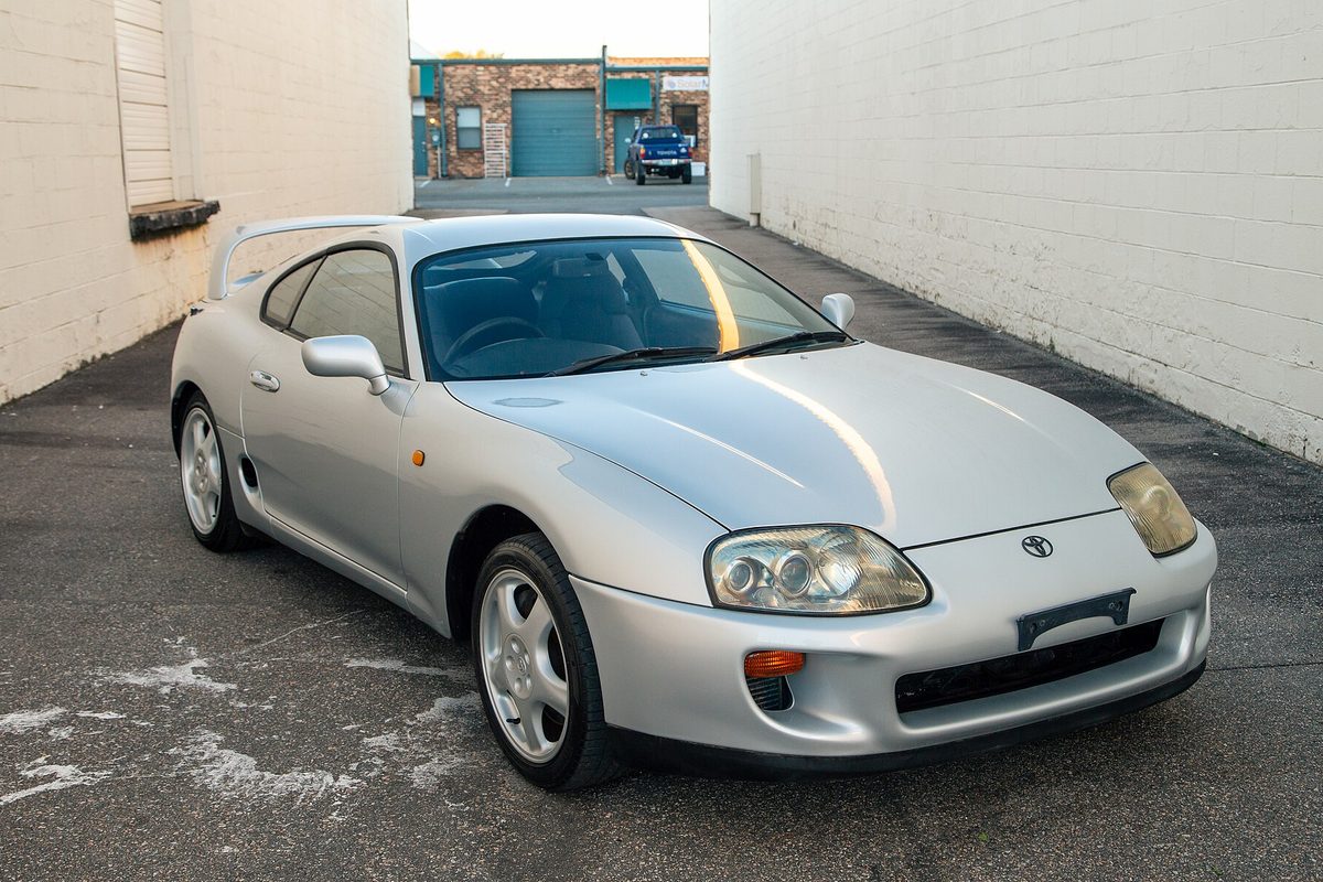 A silver fourth-generation Toyota Supra with a rear wing and silver alloy wheels, parked in a paved alley between white brick walls
