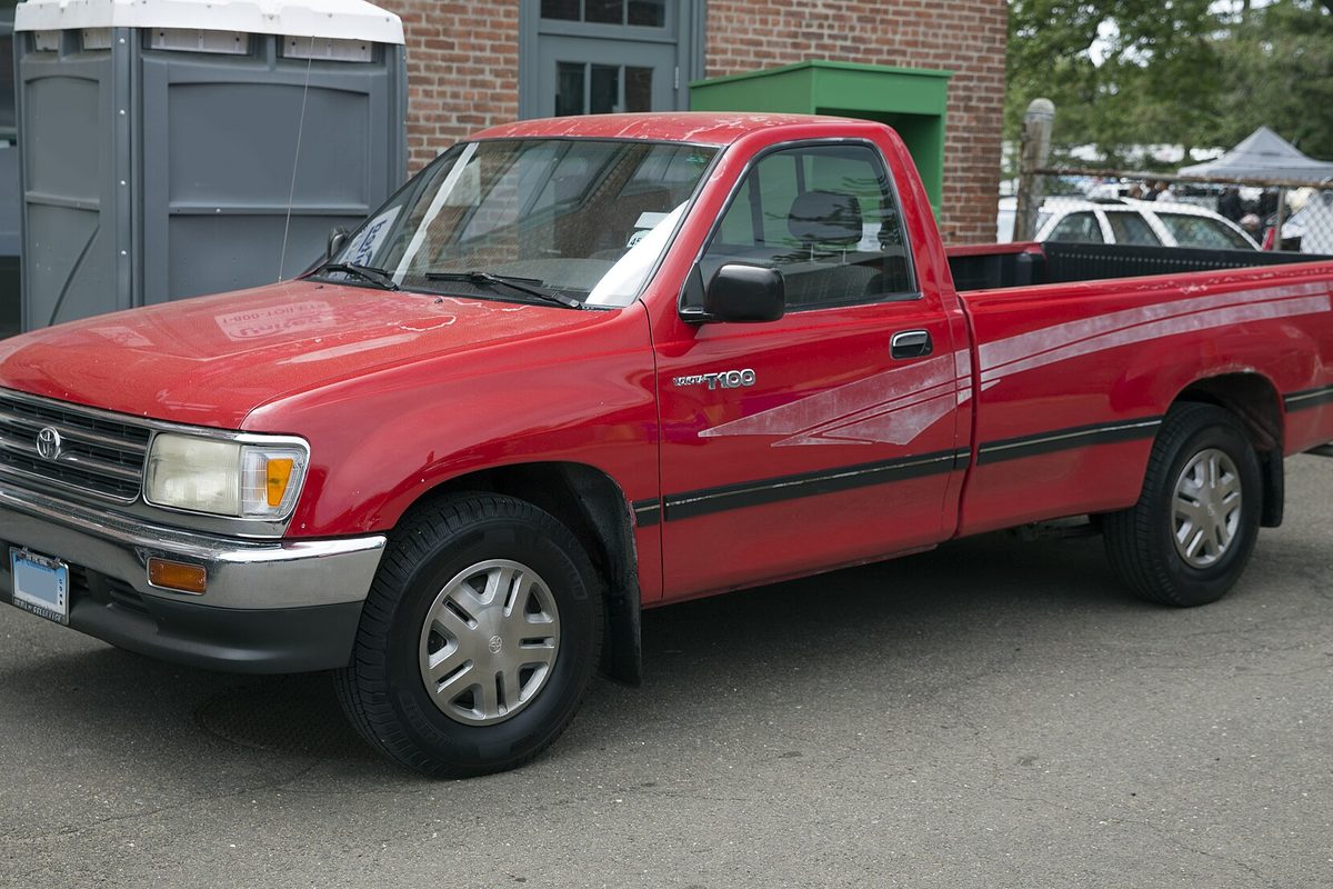 A red 1994 Toyota T100 regular-cab pickup with a long bed, stripe decals and silver wheel covers, parked beside a brick building
