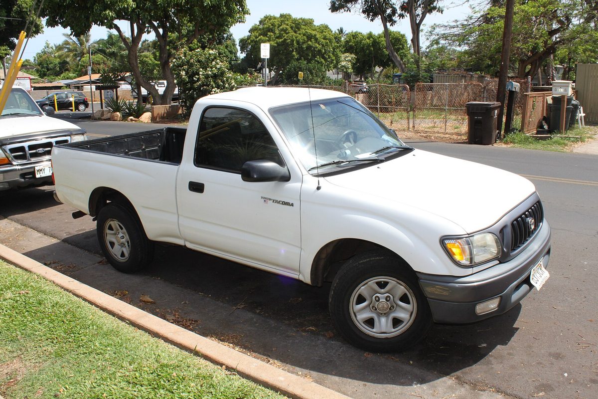 A white first-generation Toyota Tacoma regular cab with steel wheels, parked at a curb on a sunny residential street