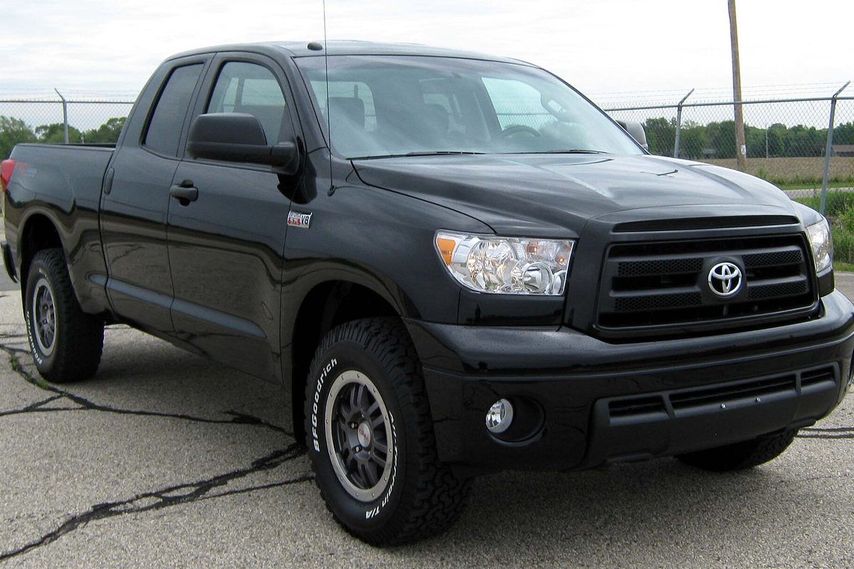 A black 2010 Toyota Tundra Double Cab on off-road tires, parked on cracked pavement in front of a chain-link fence