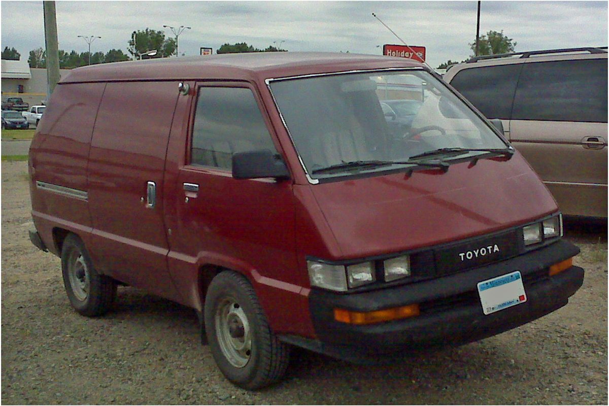 A maroon 1987 Toyota Van with blank rear side panels and steel wheels, parked on gravel beside a newer minivan