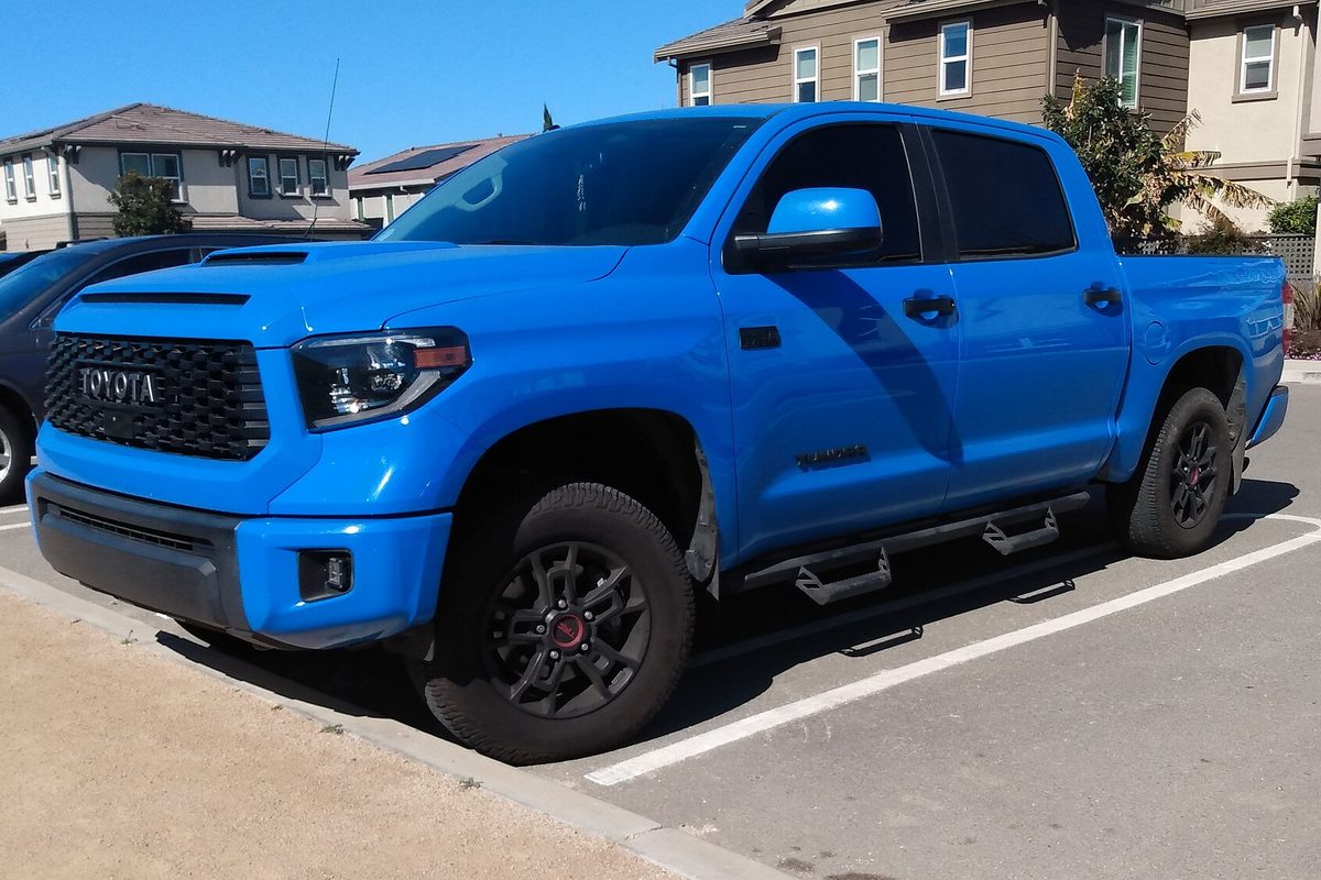 A bright blue 2019 Toyota Tundra TRD Pro crew-cab pickup with black wheels and side steps, parked in a lot in front of two-story houses