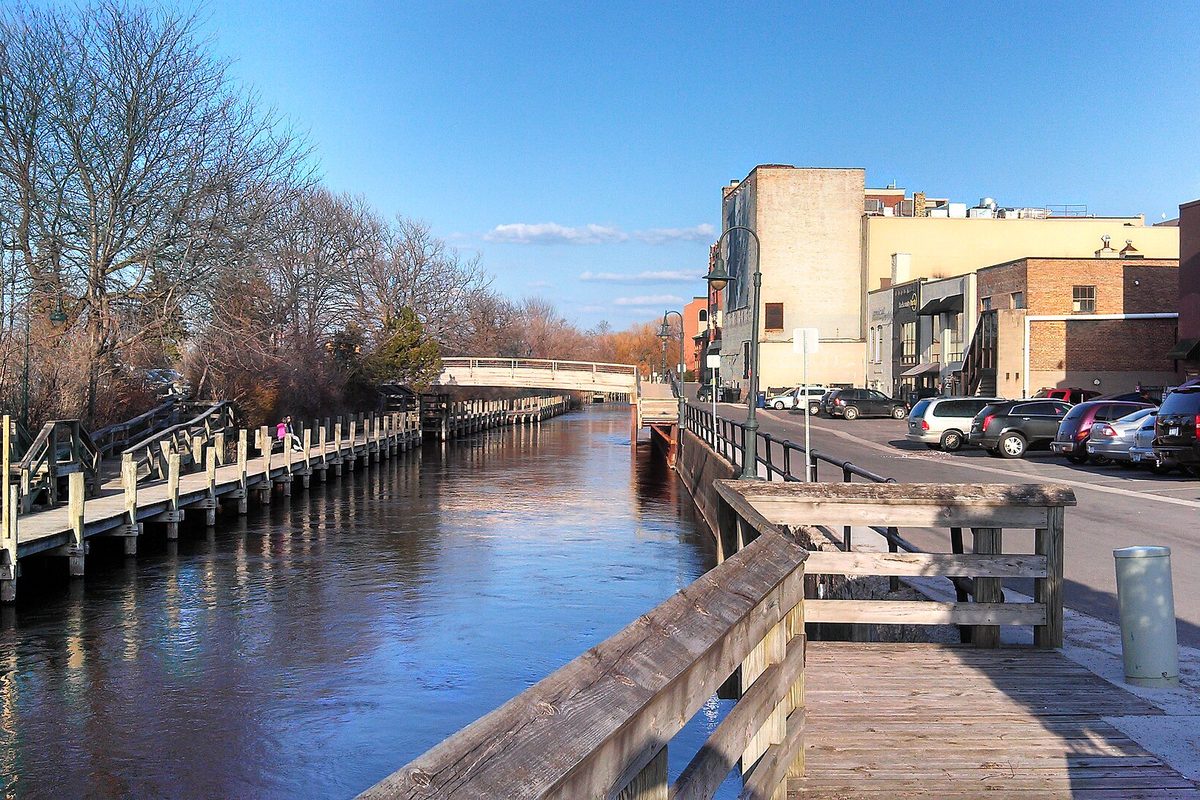 Downtown Traverse City's river walk: a wooden boardwalk beside a calm river, a footbridge, brick buildings and parked cars under a blue sky
