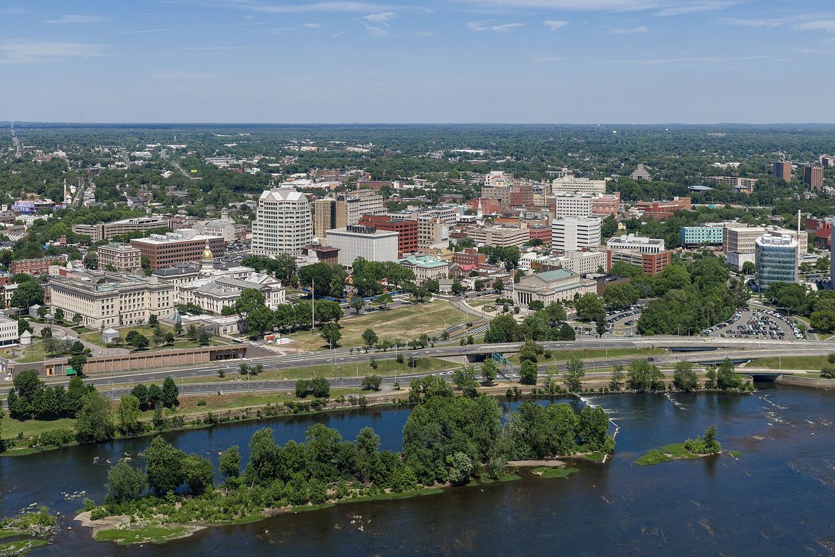 Aerial view of downtown Trenton's office buildings and a gold dome, with a wide river and wooded islands in the foreground