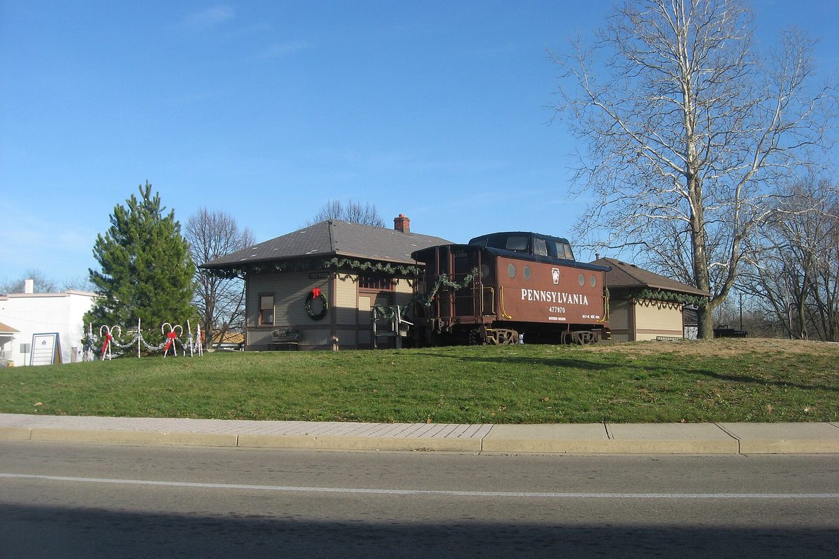 The small 1913 Trotwood railroad station with a red Pennsylvania caboose beside it, decked with holiday wreaths, on a grassy rise above Main Street