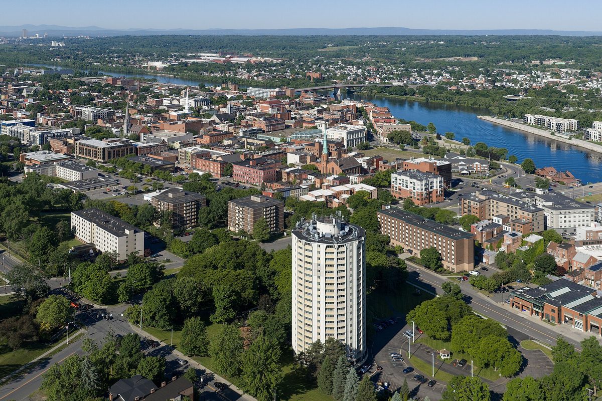 Aerial view of Troy with a round white apartment tower in front and the Hudson River curving past downtown