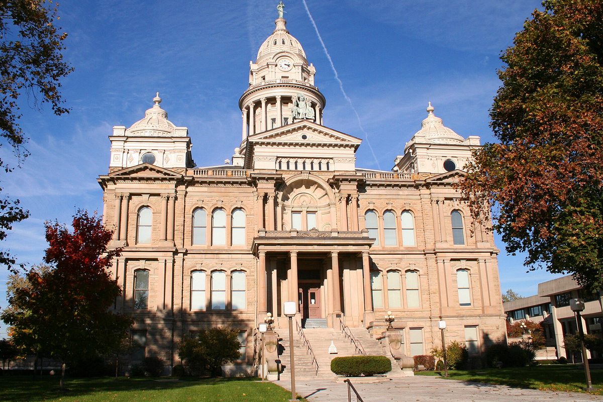 The domed tan stone courthouse in Troy, Ohio, with red and orange autumn trees on both sides and a blue sky behind