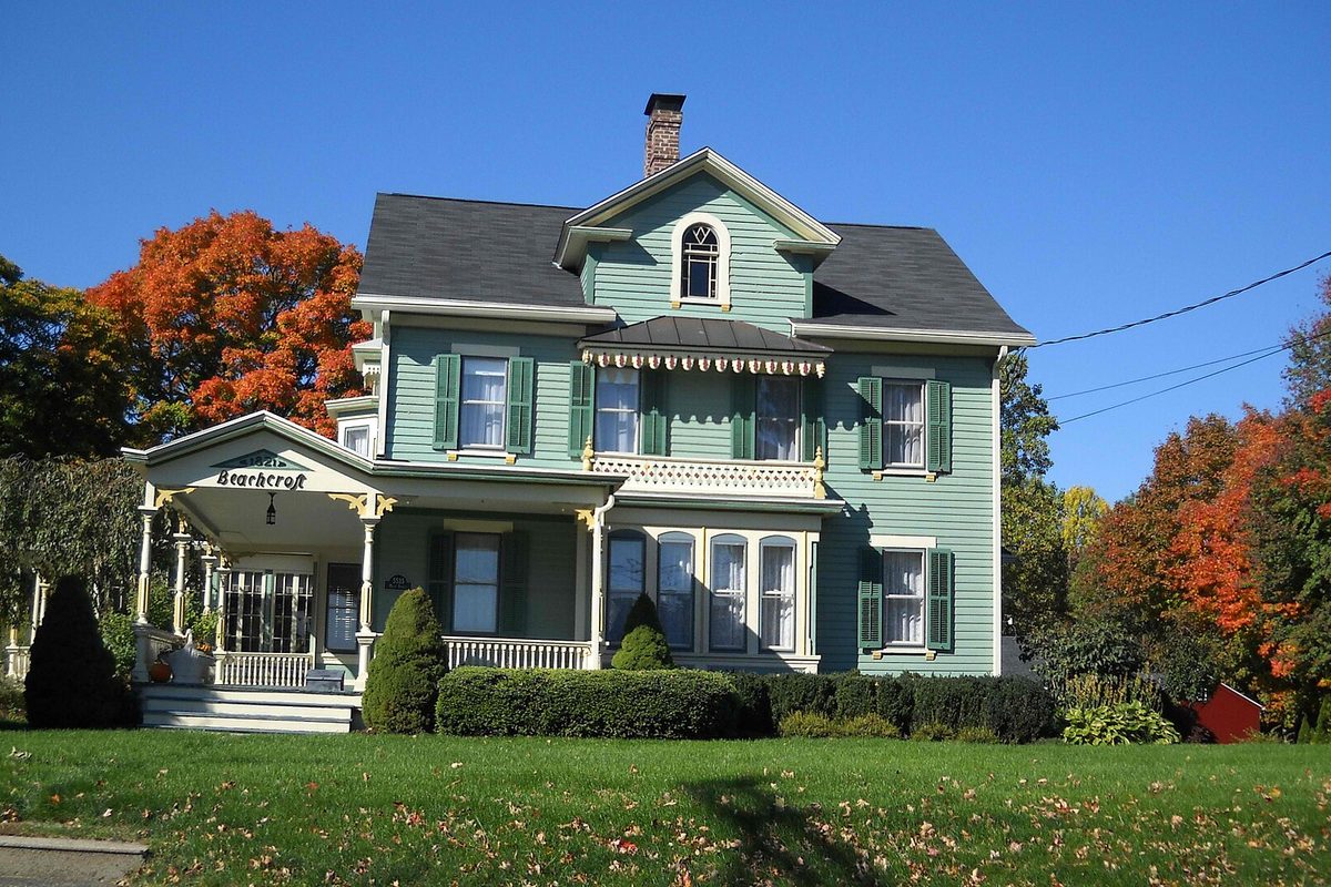The green 1821 Beachcroft house on Main Street in Trumbull, with a white front porch, under blue sky and orange autumn trees