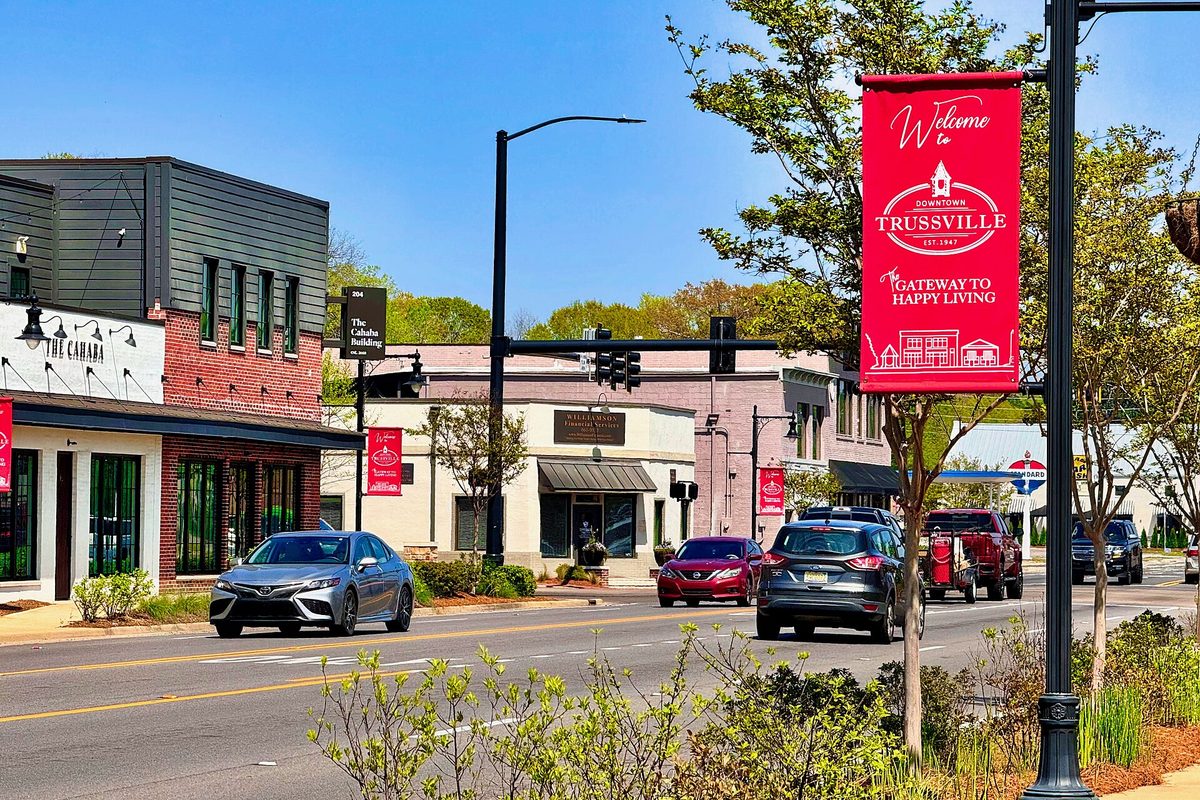 A red 'Welcome to Downtown Trussville' banner on a black lamppost over Main Street, with cars passing brick and white storefronts under a blue sky