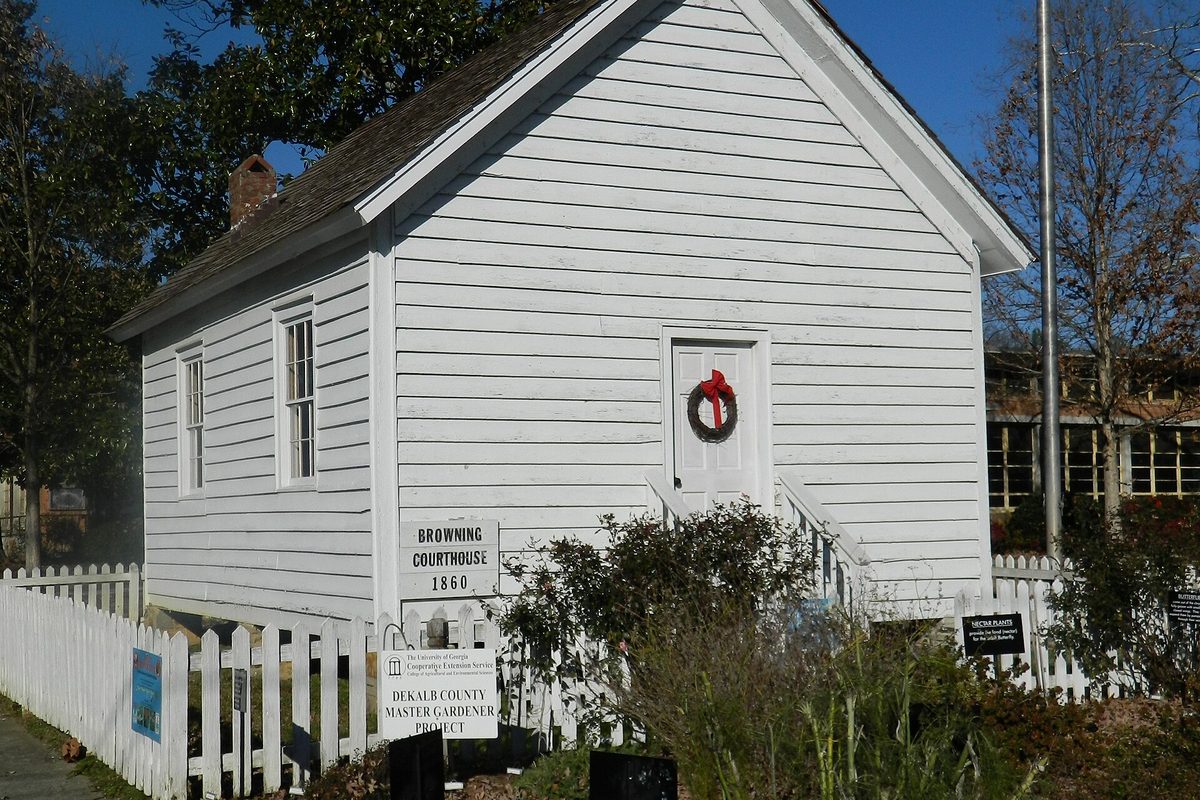 The white clapboard Browning Courthouse in Tucker, built in 1860, behind a picket fence with a wreath on its door and a DeKalb County Master Gardener sign