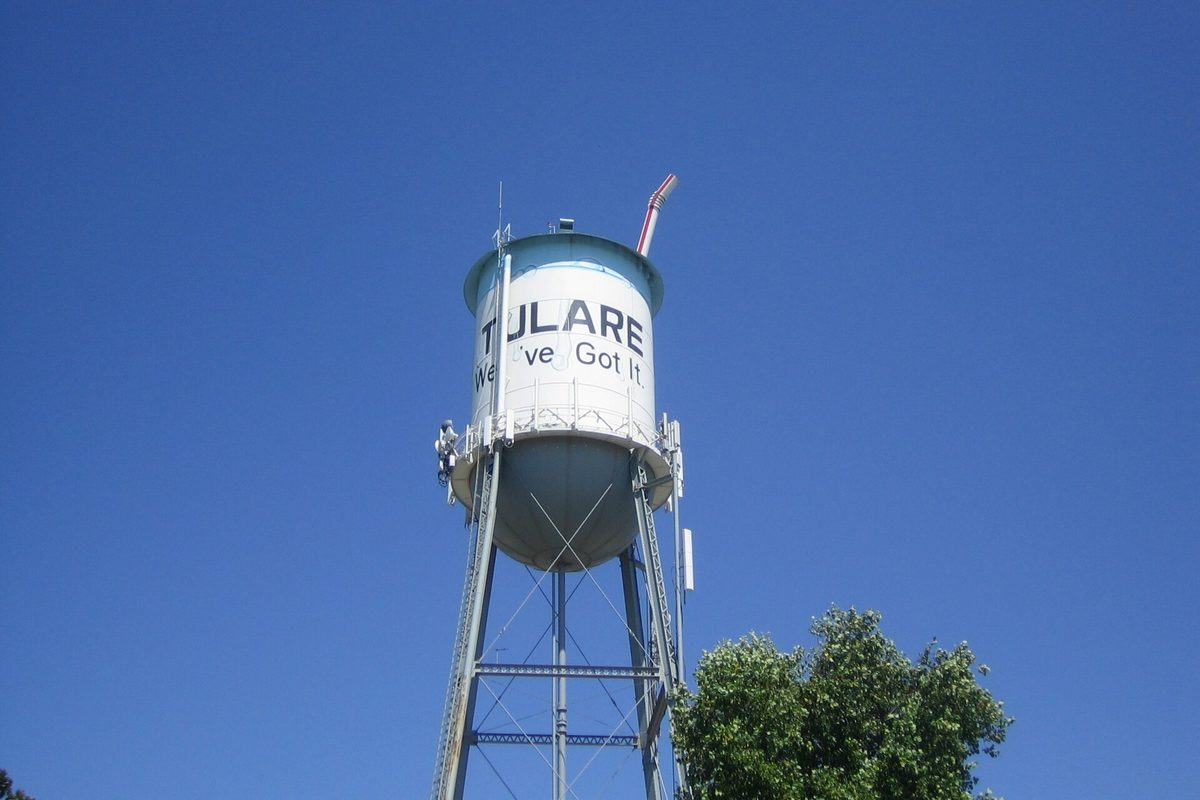 Tulare's white water tower reading 'Tulare, We've Got It' against a deep blue sky, with a treetop at the lower right