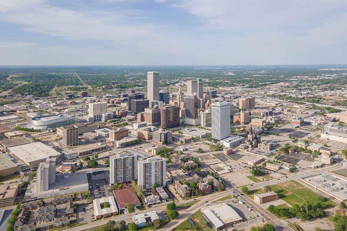 Aerial view of downtown Tulsa's high-rise towers, with a round arena at left and low neighborhoods and trees stretching to the horizon