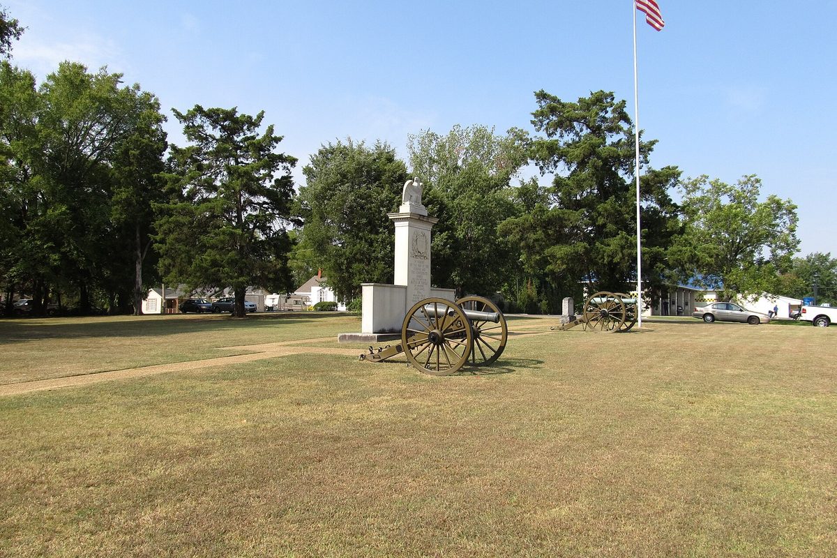 A white stone monument between two old cannons on wooden spoked wheels on a mowed field at Tupelo National Battlefield, with a flagpole and trees behind