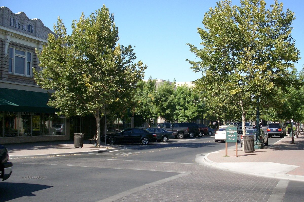 Tree-lined Main Street in downtown Turlock, with cars parked along the curb and an old two-story brick commercial building on the left