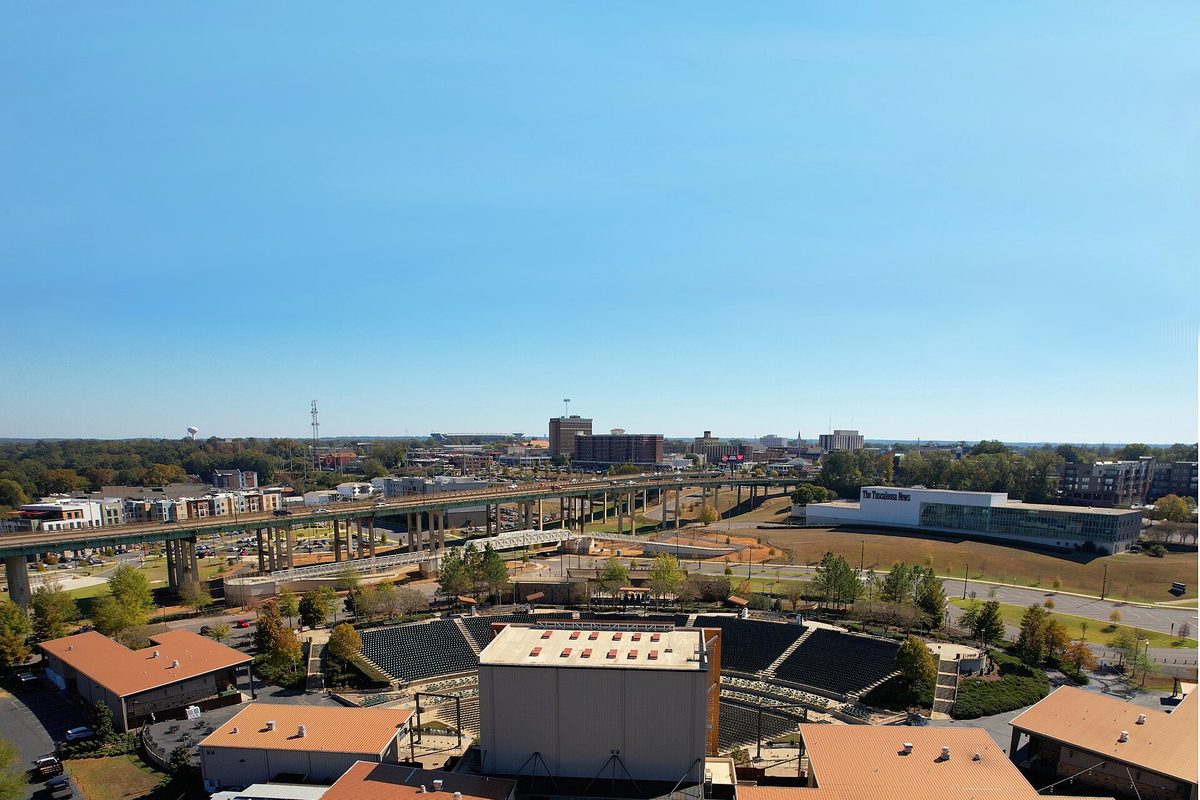 Downtown Tuscaloosa from above on a clear day, with an outdoor amphitheater in front, a long highway bridge behind it and the skyline beyond