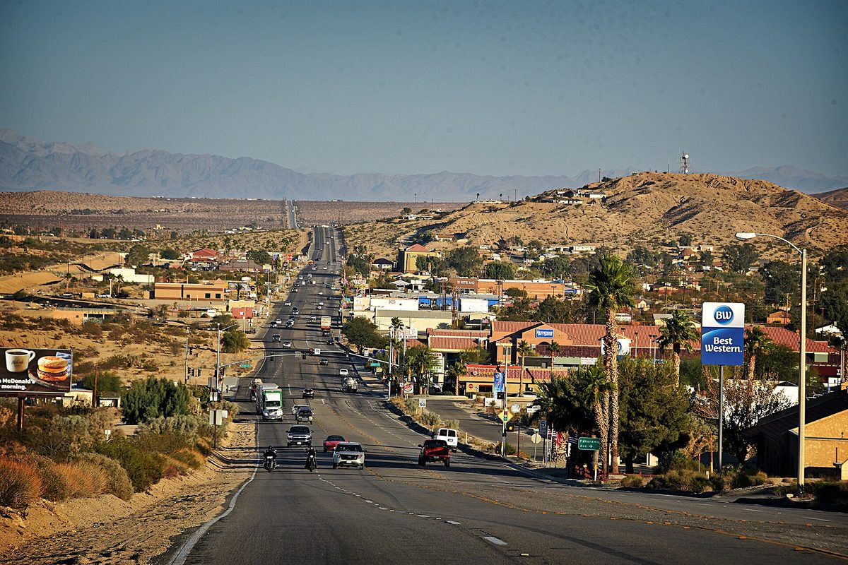 Highway 62 running east through Twentynine Palms past palm trees, low buildings and a motel sign, with dry hills and distant mountains behind