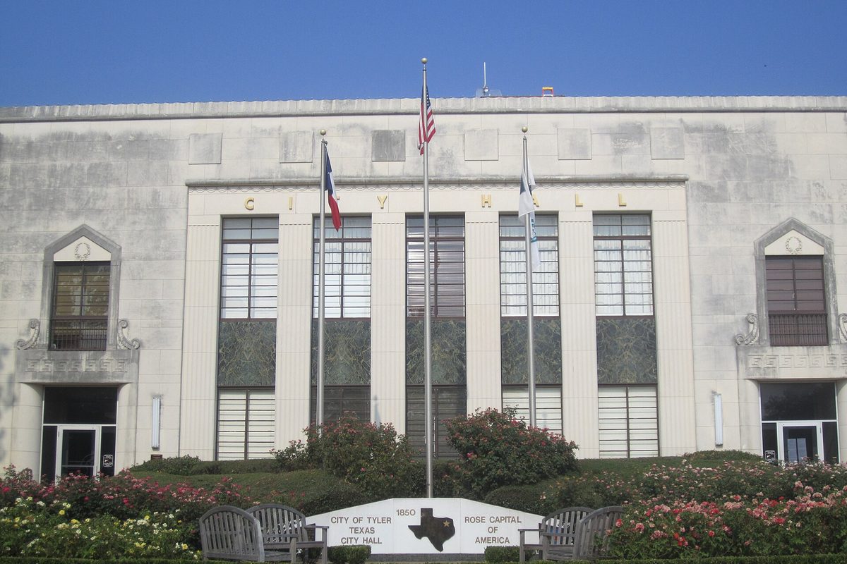 Tyler City Hall, a pale stone building with tall vertical windows and three flags, behind rose bushes and a sign reading City of Tyler Texas City Hall, Rose Capital of America