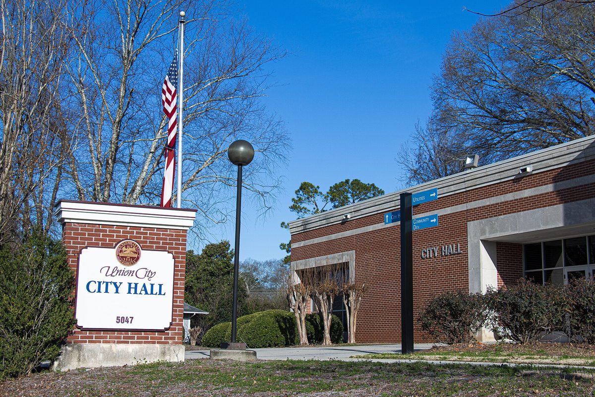 A brick sign reading Union City City Hall 5047 beside a flagpole, in front of the low red-brick City Hall building under a clear blue sky