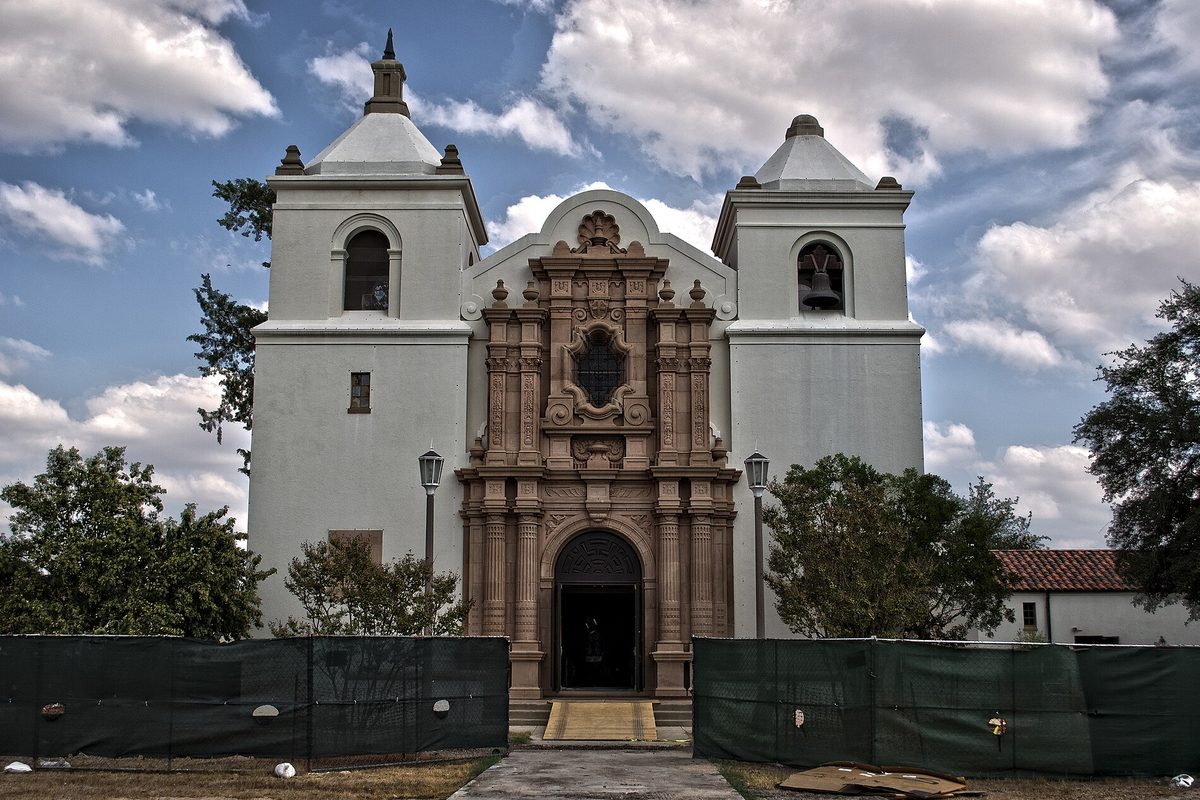 A white Spanish mission-style church with two bell towers and a carved stone doorway, behind green construction screening on a cloudy day